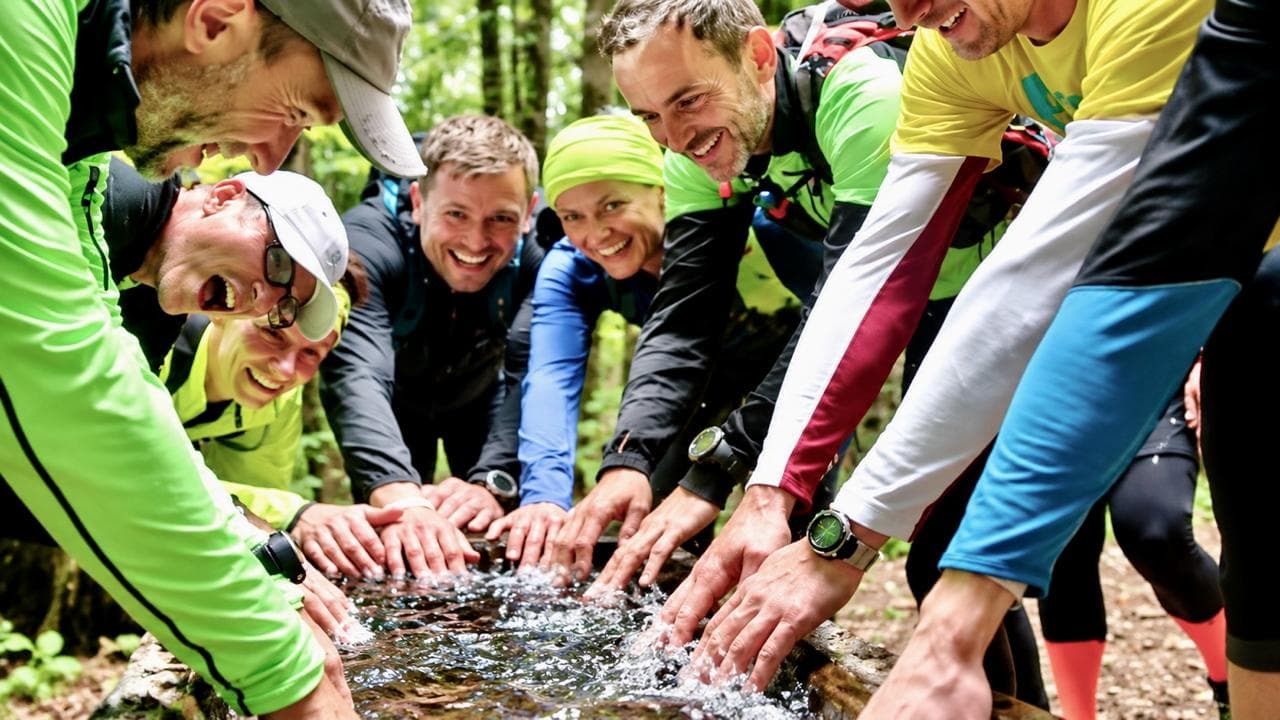 Pavel Hruška s běžeckým týmem v lese, všichni se smějí a společně namáčejí ruce do studánky